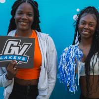 two people posing in front of CAB backdrop at Laker Kickoff photo booth and holding GV sign and blue and white pom poms